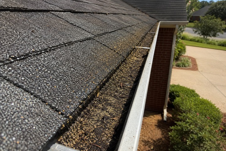 Dark gritty sludge and shingle granules filling a gutter beside weathered asphalt shingles on a Georgia brick home