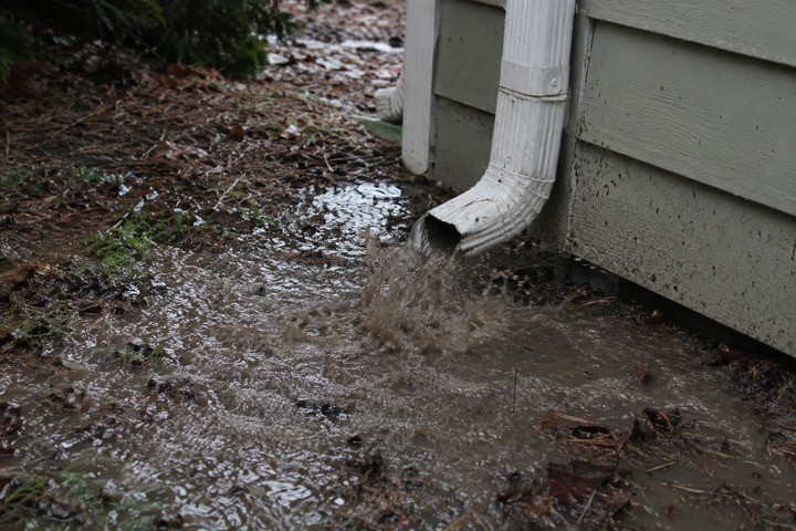 White downspout discharging muddy stormwater that pools and erodes the ground at the foundation of a Georgia home